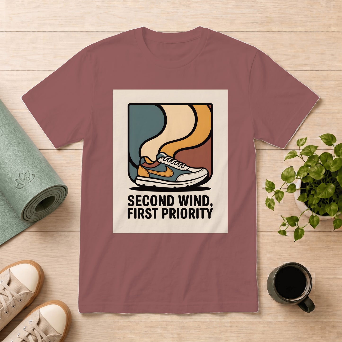 A mauve short-sleeve graphic T-shirt laid flat on a light wood surface, surrounded by a rolled yoga mat, sneakers, a potted plant, and a coffee mug. The shirt has a large rectangular front print showing a stylized sneaker illustration with the text 'SECOND WIND, FIRST PRIORITY'.