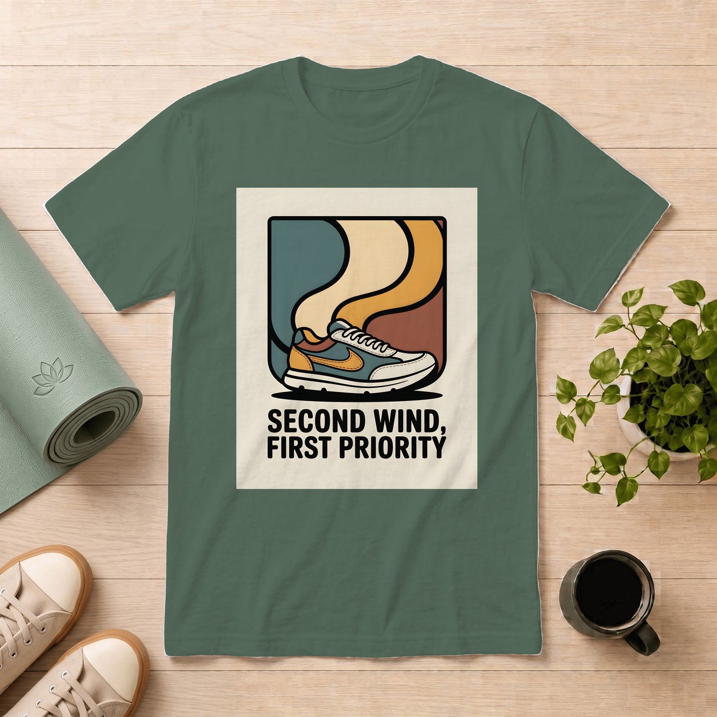 A sage green short-sleeve t-shirt with a retro graphic of a sneaker and the text 'SECOND WIND, FIRST PRIORITY' in black, displayed in a flat-lay arrangement on a light wood background with a yoga mat, shoes, a plant, and a coffee mug.
