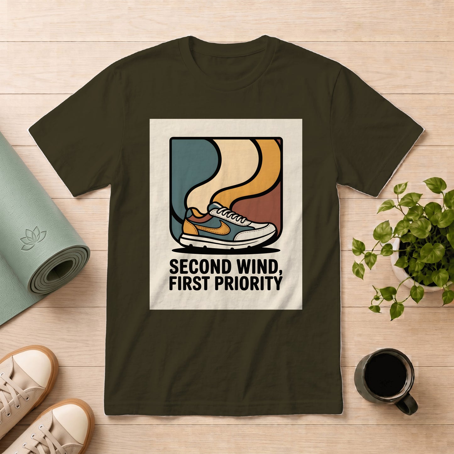 An olive green t-shirt with a retro graphic of a sneaker and the text 'SECOND WIND, FIRST PRIORITY' in black. The shirt is displayed in a flat-lay arrangement on a light wood background, surrounded by a rolled yoga mat, a potted plant, a coffee mug, and sneakers.