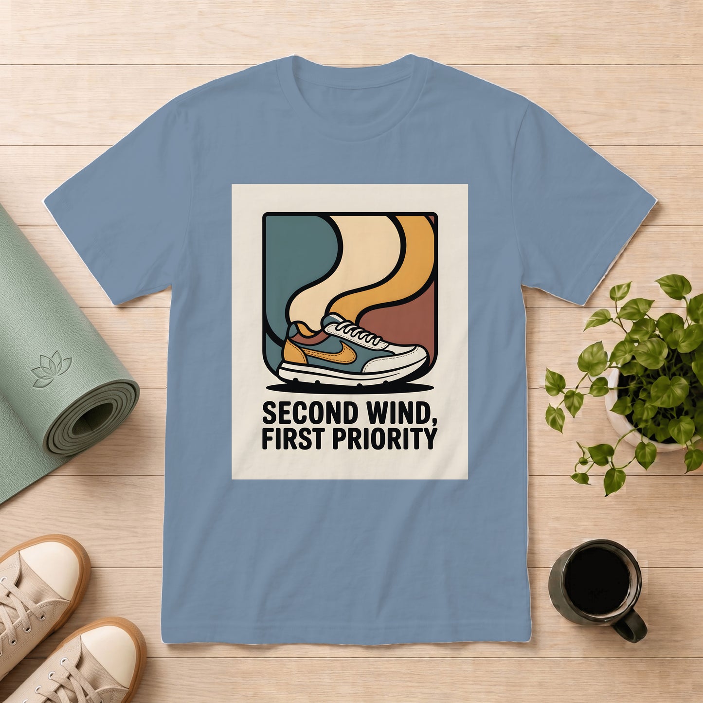 A light blue crewneck t-shirt laid flat on a light wood surface, featuring a retro graphic of a sneaker with the text 'SECOND WIND, FIRST PRIORITY'. The shirt is styled with a rolled green yoga mat, beige sneakers, a potted plant, and a coffee mug.