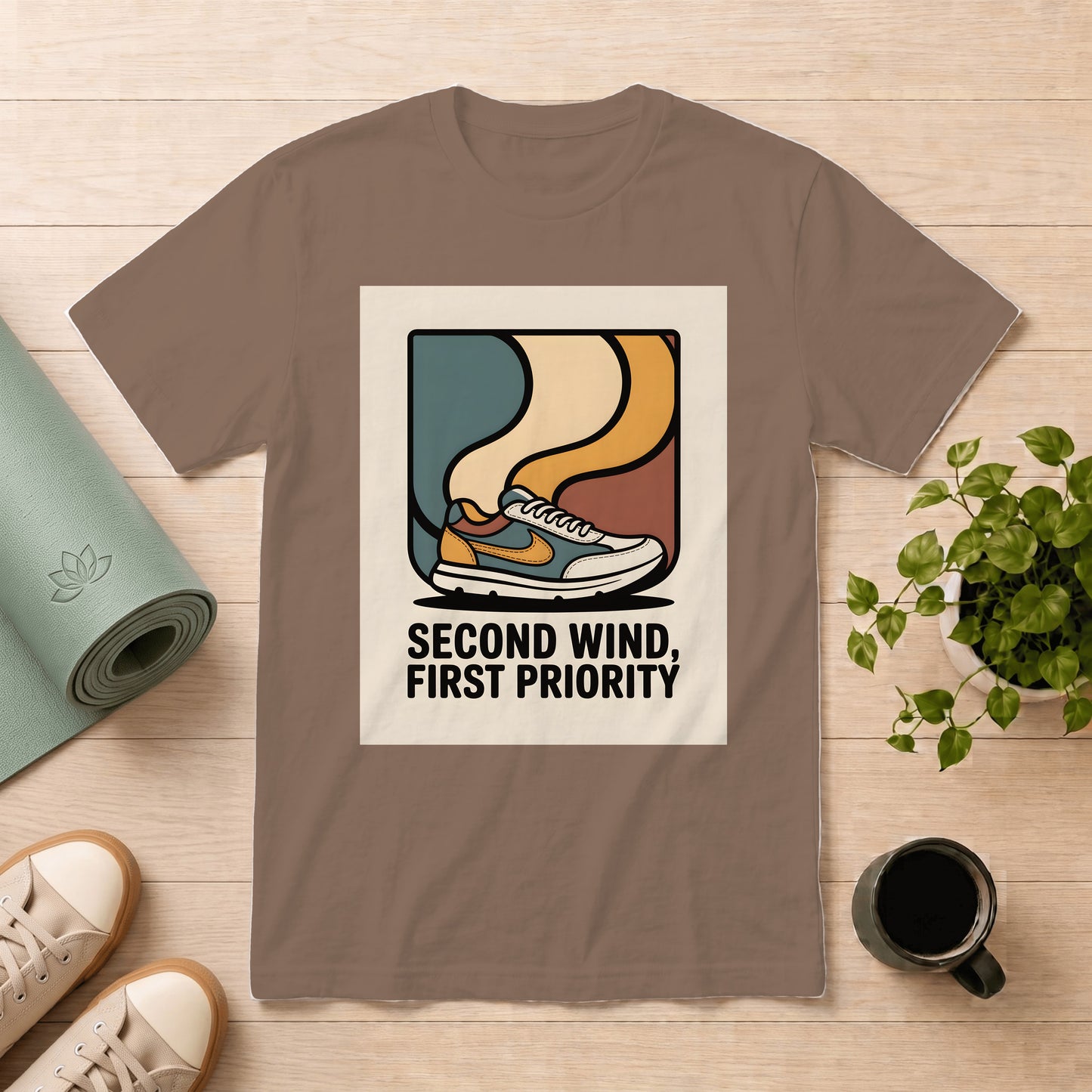 A taupe brown short-sleeve t-shirt with a retro graphic of a sneaker and the text 'SECOND WIND, FIRST PRIORITY' on the front, displayed in a flat-lay arrangement on a light wood background with a yoga mat, shoes, a plant, and a coffee mug.